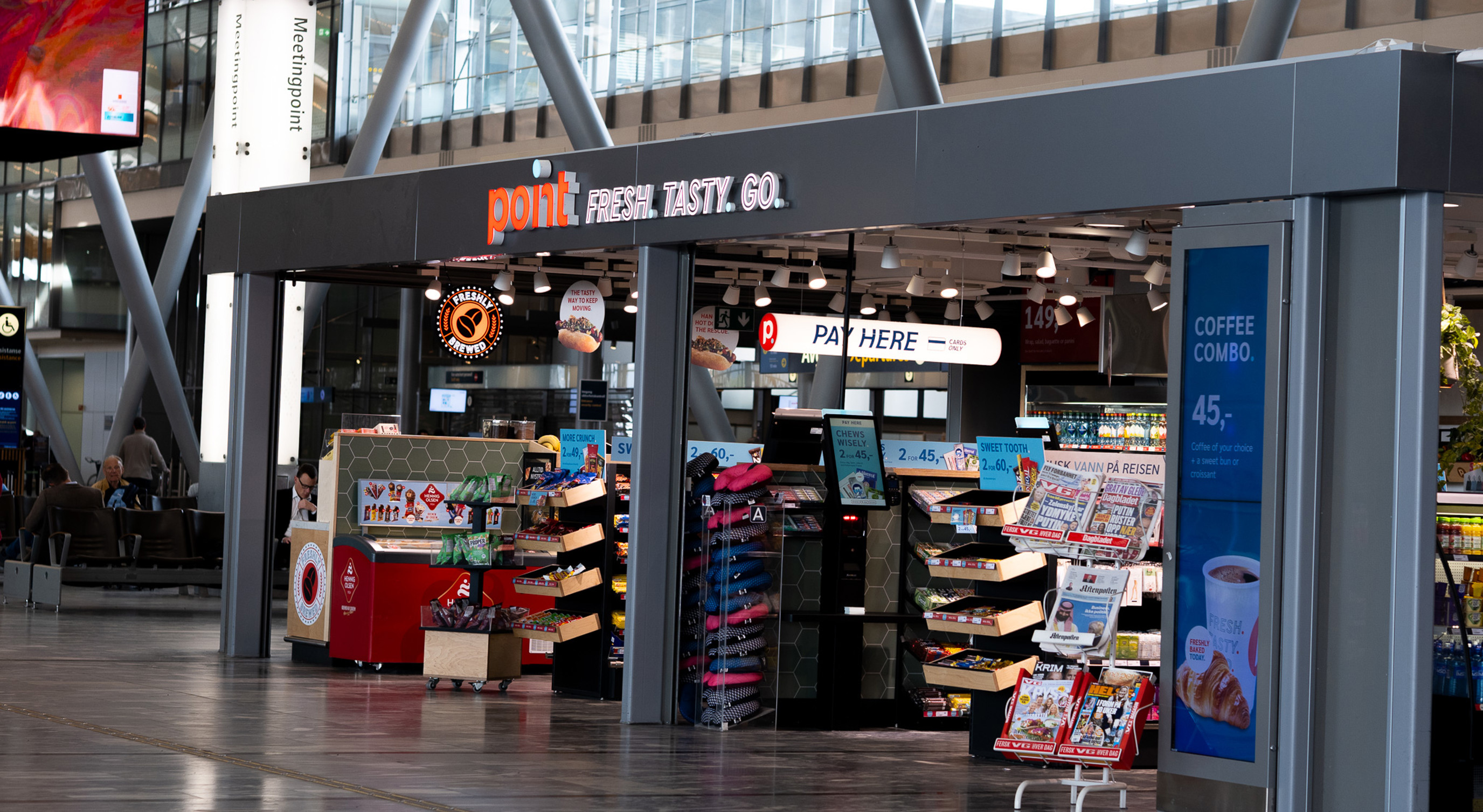 Butikk ved flyplass, med skilting "point" og "fresh. tasty. go." sel ulike varer som aviser, snacks og drikke. Reklametavle for kaffi. Moderne interiør med reisande i bakgrunnen