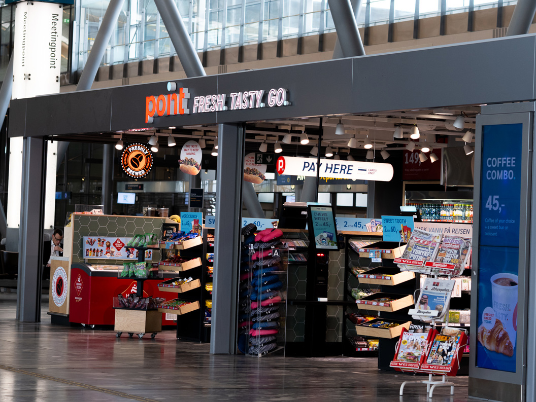 Butikk ved flyplass, med skilting "point" og "fresh. tasty. go." sel ulike varer som aviser, snacks og drikke. Reklametavle for kaffi. Moderne interiør med reisande i bakgrunnen