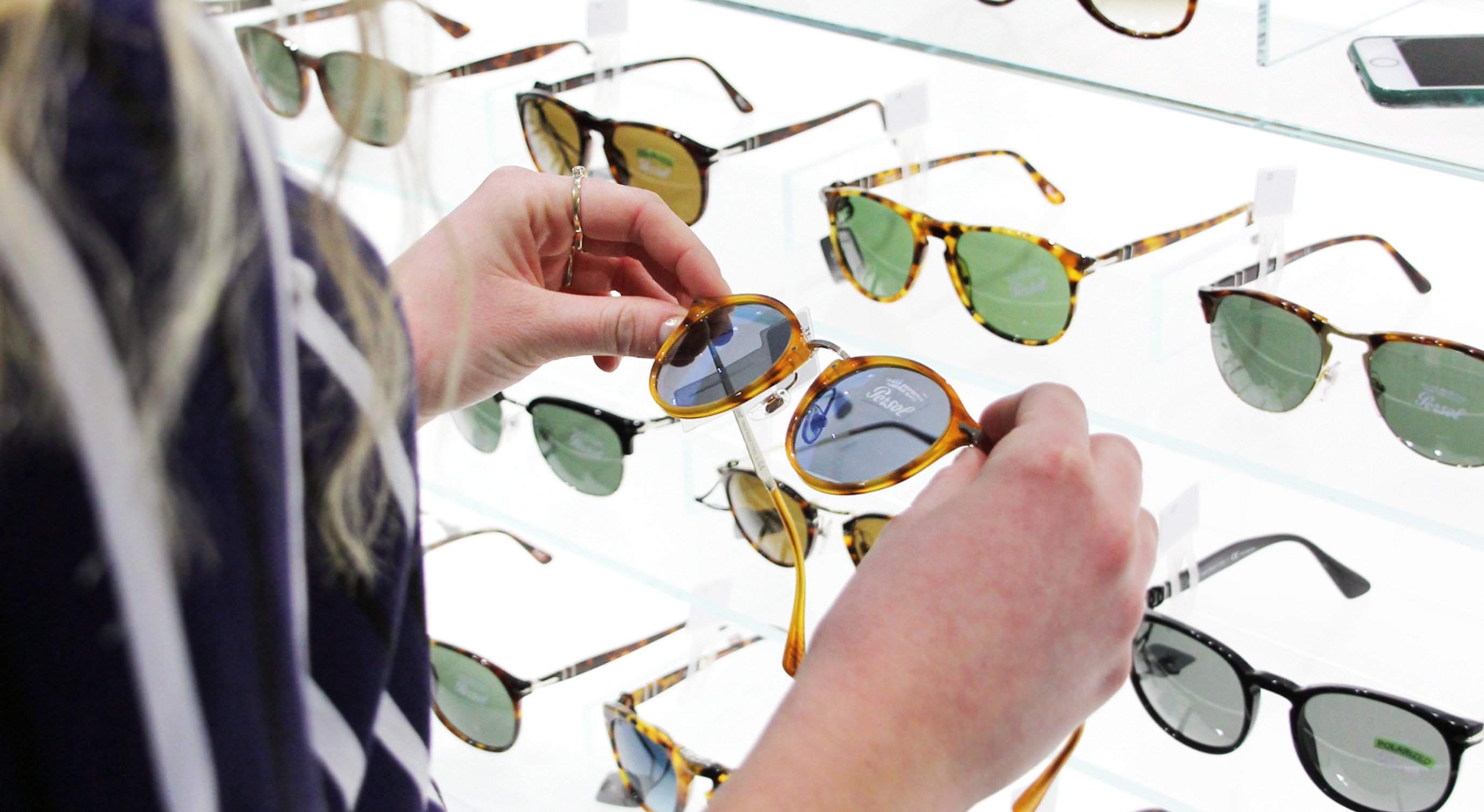A person examining a selection of sunglasses in a retail display, with various styles and colors, at a store counter