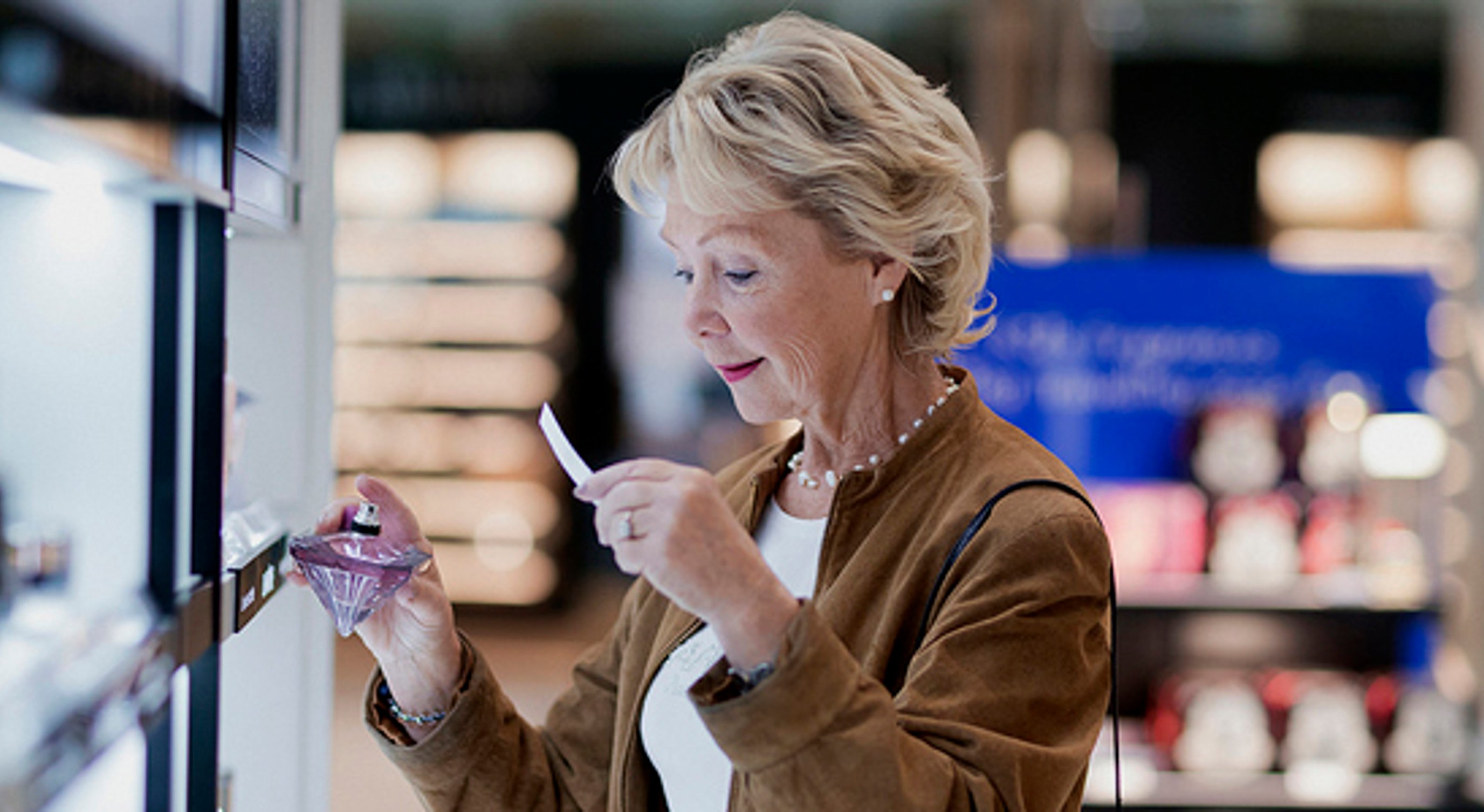 Senior woman testing perfume scent in a store, holding a fragrance bottle and sample paper strip, with shelves of products in the background