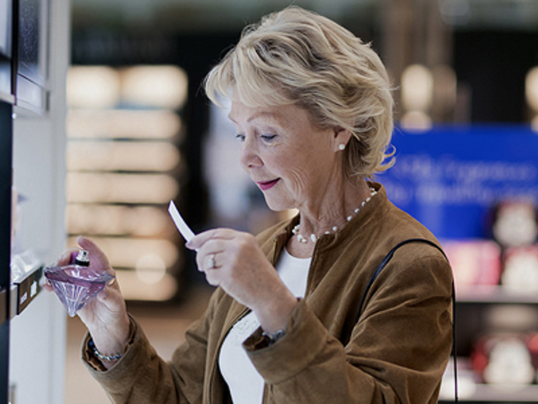 Senior woman testing perfume scent in a store, holding a fragrance bottle and sample paper strip, with shelves of products in the background