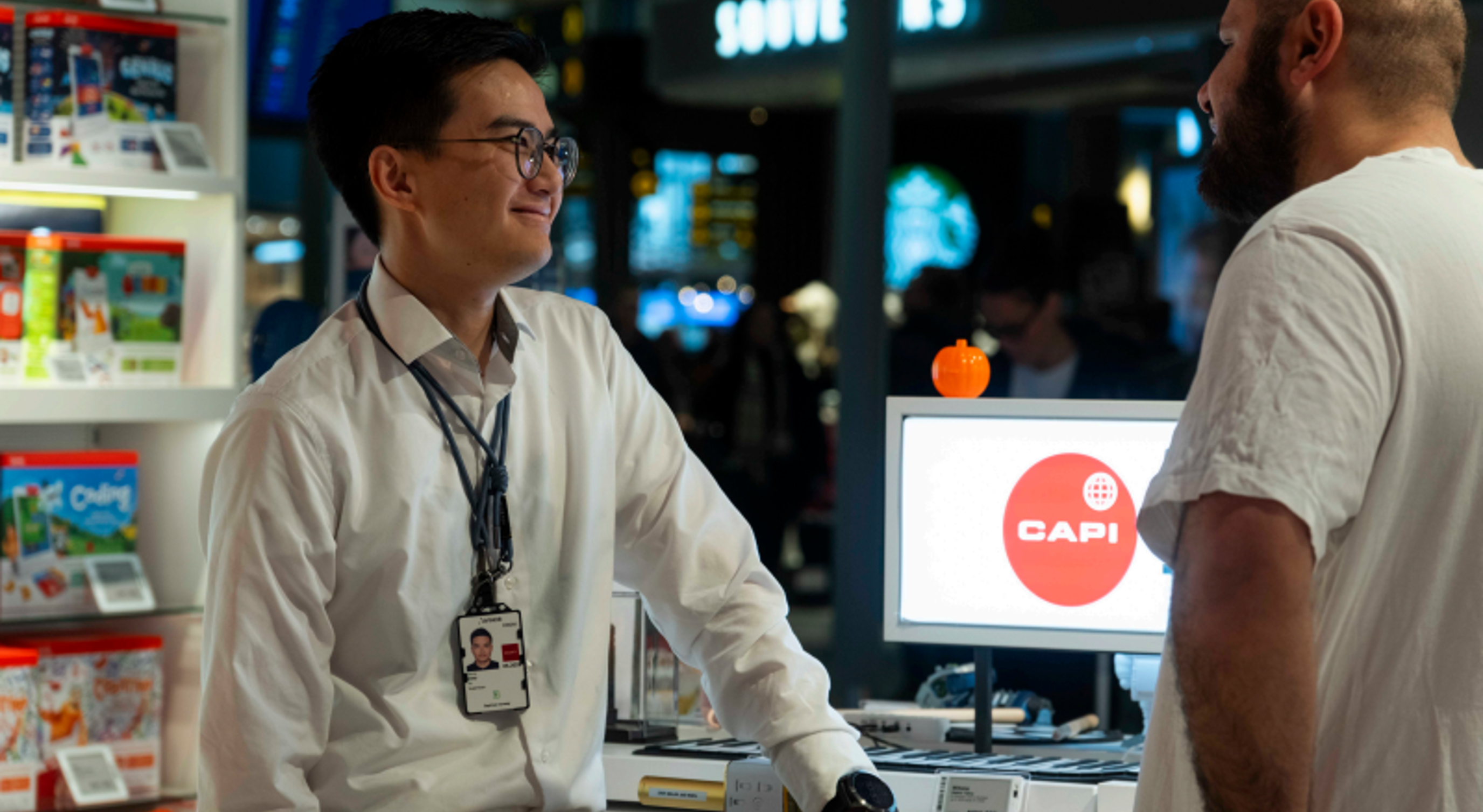 Butikkmedarbeidar møter ein kunde ved skranken i ein elektronikkbutikk, med produktutstilling og ein dataskjerm med Capi-logo i bakgrunnen