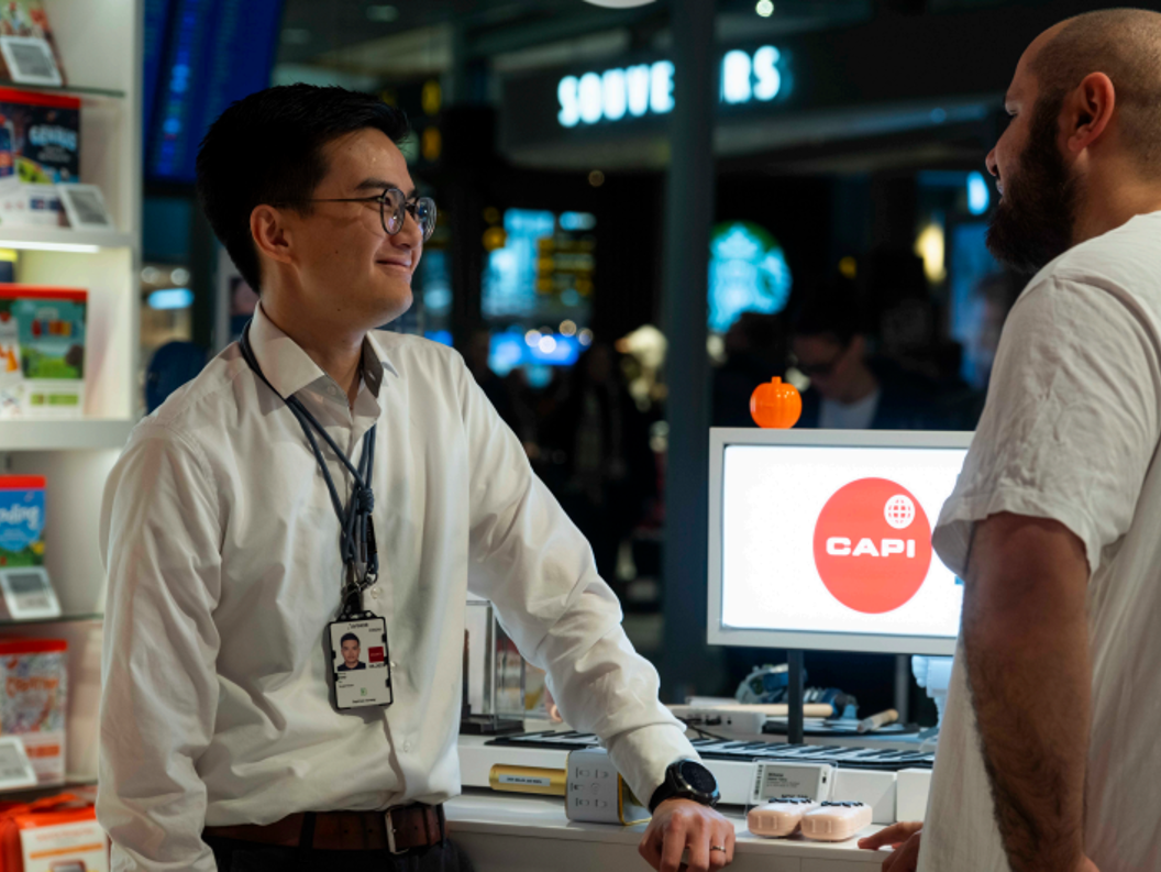 Butikkarbeidar i kvit skjorte hjelper ein kunde ved ein elektronikkdisk i Capi-butikken på ein flyplass