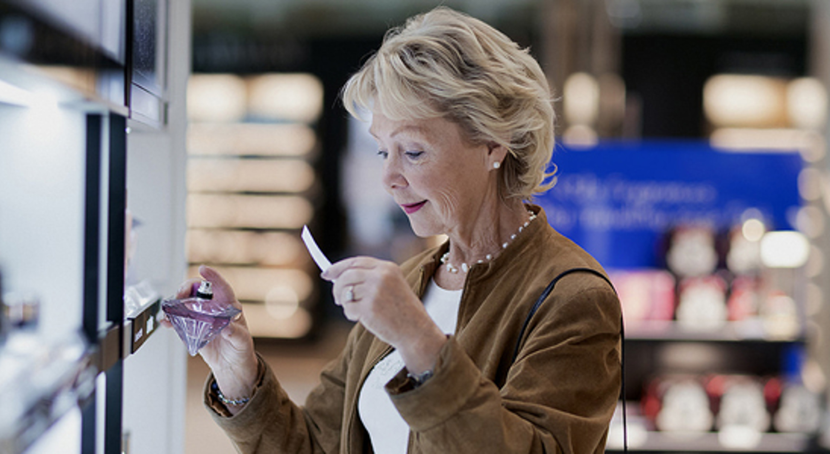 Older woman testing perfume at a store counter, holding a fragrance bottle and test strip, with shelves of products in the background