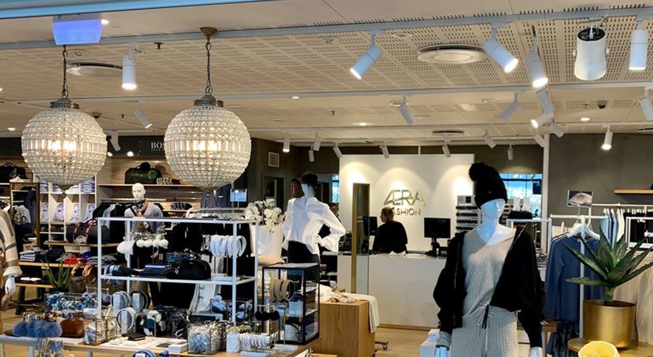 Fashion retail store entrance showcasing stylish mannequins and accessories, with prominent "AERA Fashion" signage above. The interior displays a variety of clothing and jewelry under warm lighting