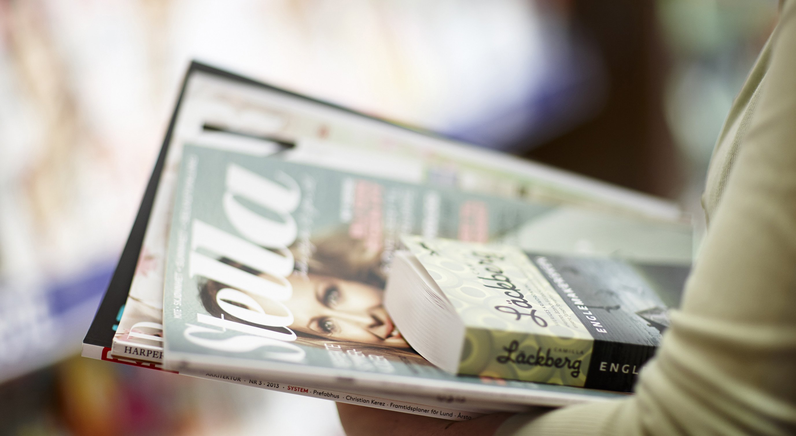 Person holding a stack of magazines and books, featuring a visible "Stella" magazine on top, with a blurred background in a bookstore setting