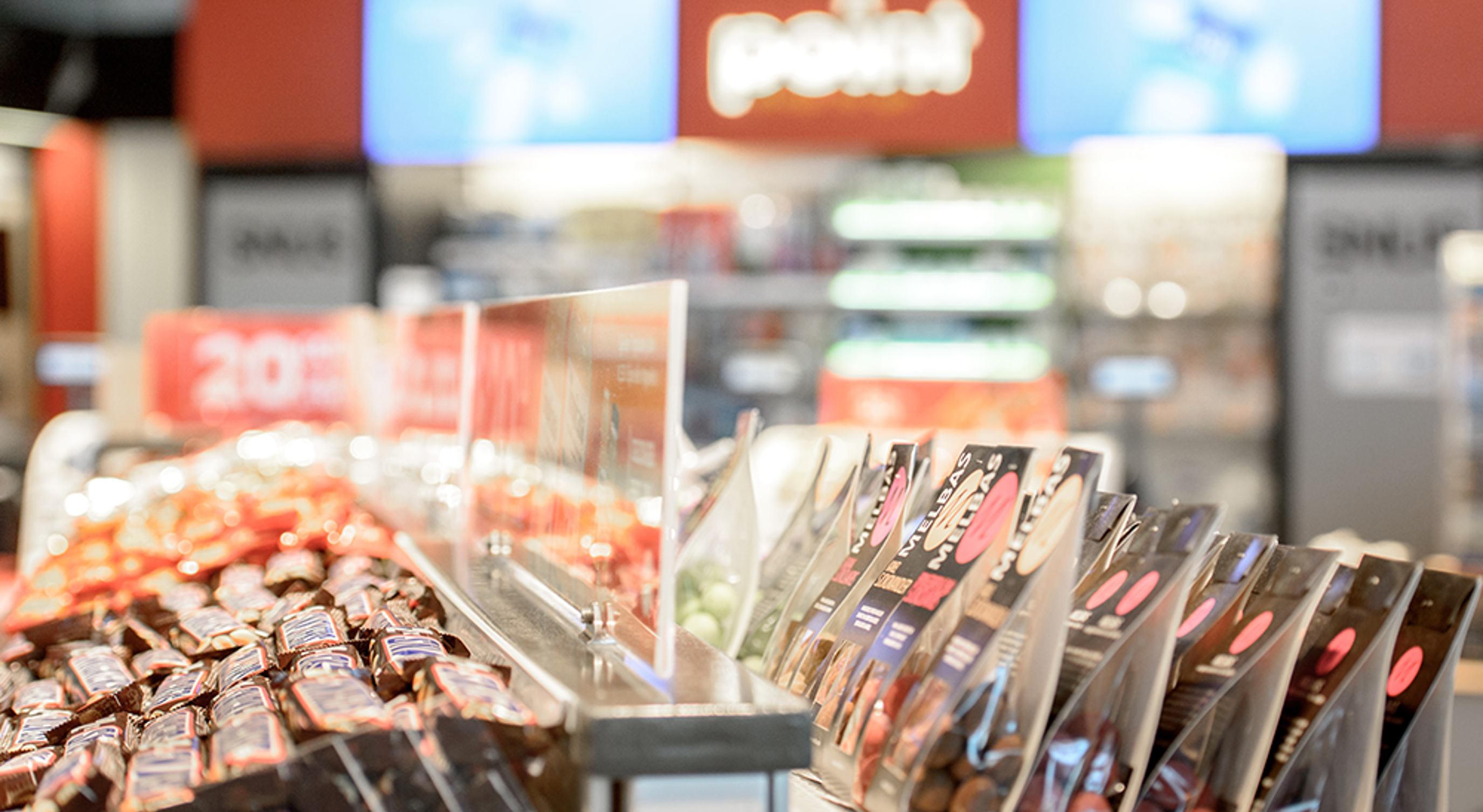 Snack display in a convenience store with various packaged candies and chocolates, featuring promotional signs and a beverage cooler in the blurred background