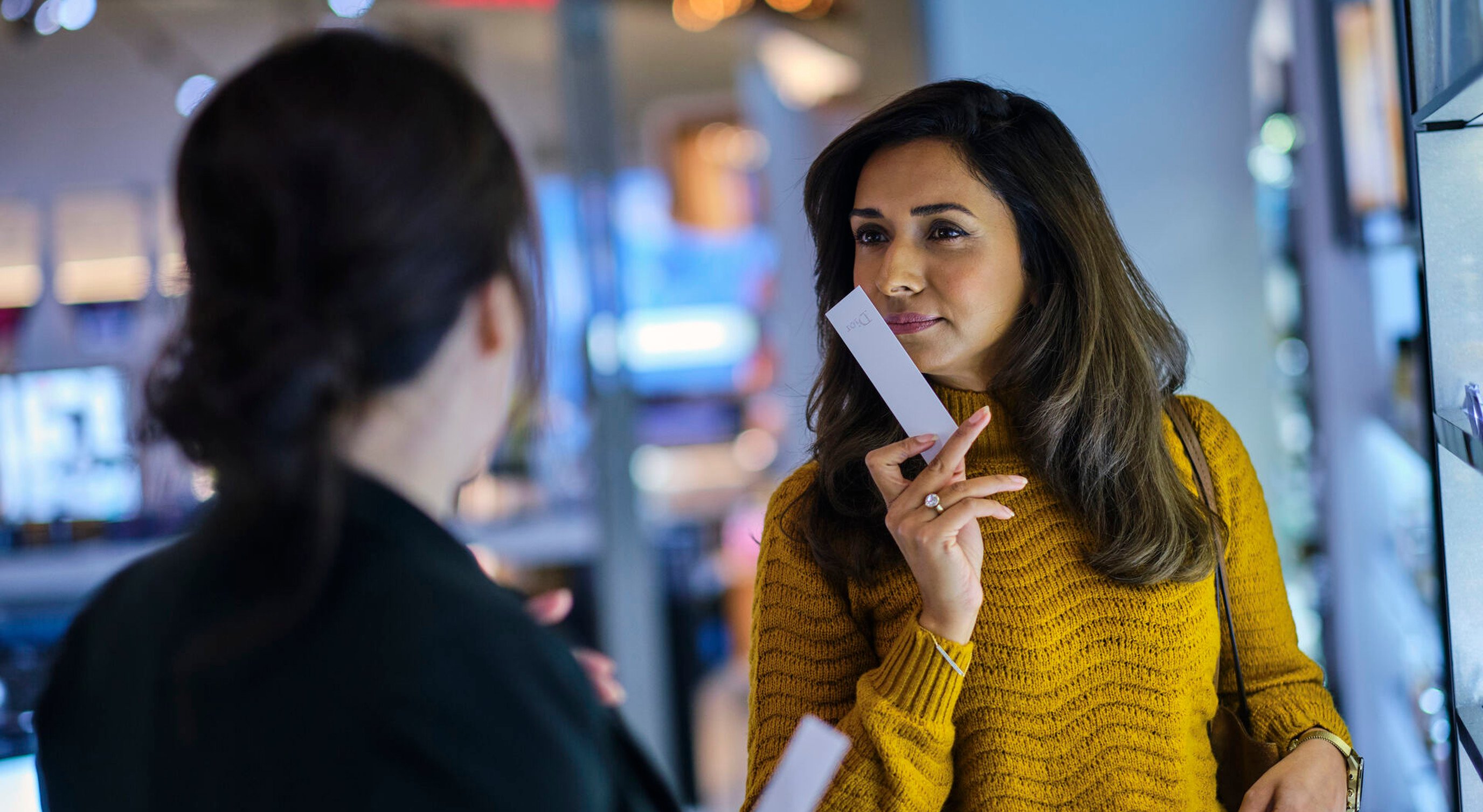 Kvinne i gul genser tester parfyme i en butikk, holder en duftstrimmel og snakker med en ansatt