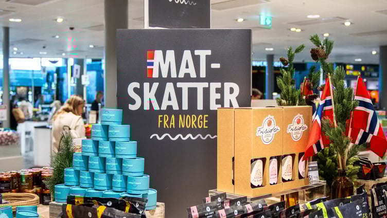 Norsk matutstilling i en butikk som viser et utvalg av pakkede varer, inkludert müsli og syltetøy, med skilt merket "Mat-Skatter fra Norge" og små norske flagg til dekorasjon