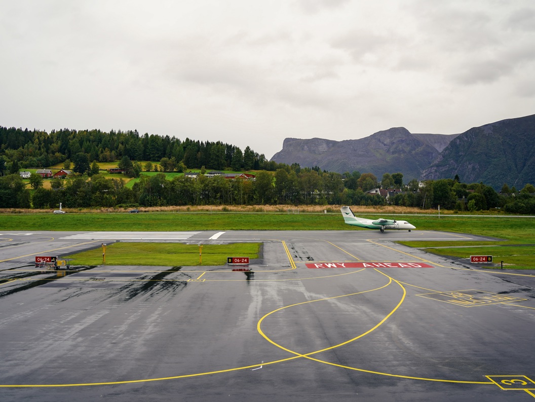 Airport landscape with a small passenger plane on the runway, surrounded by green hills and mountains in the background under a cloudy sky.