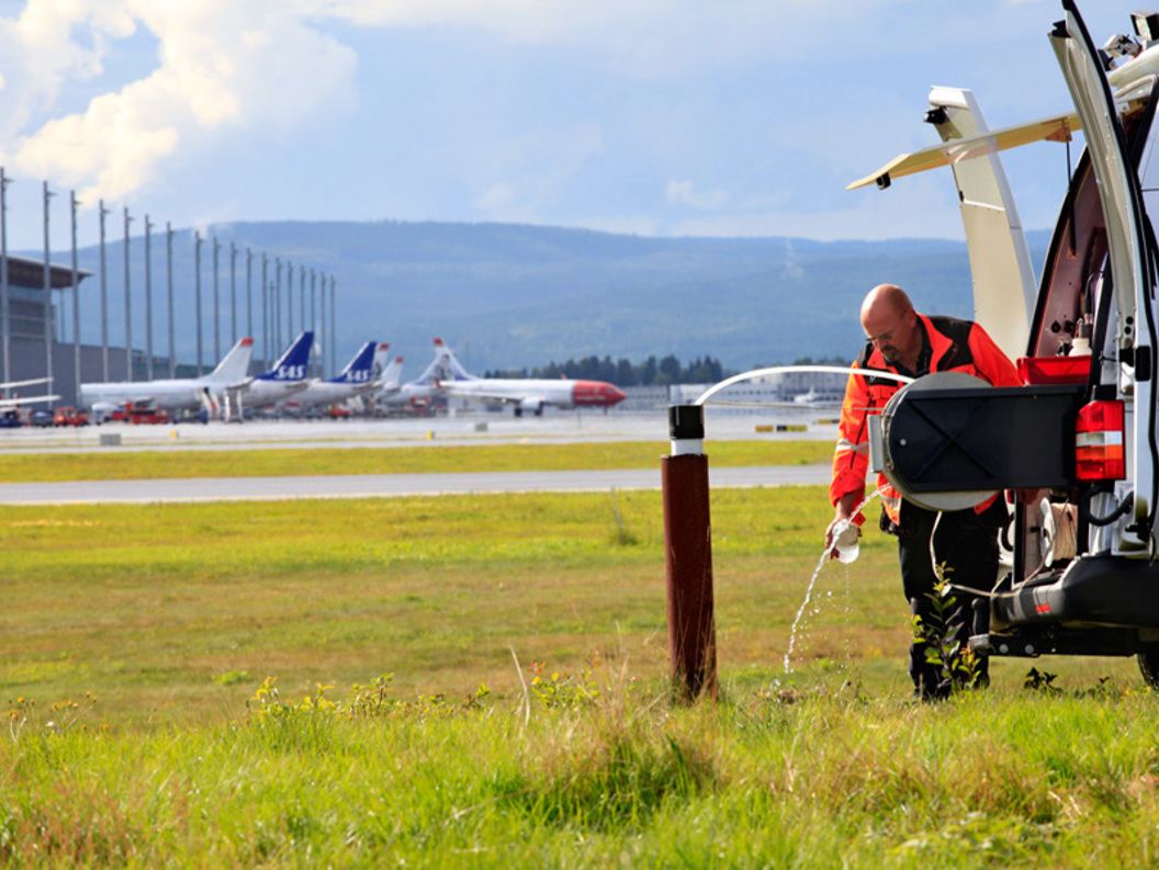 Mann i arbeidsklede fyller væske frå ein kvit varebil ved ein grassplen nær ein flyplass, med fleire fly på bakken i bakgrunnen.