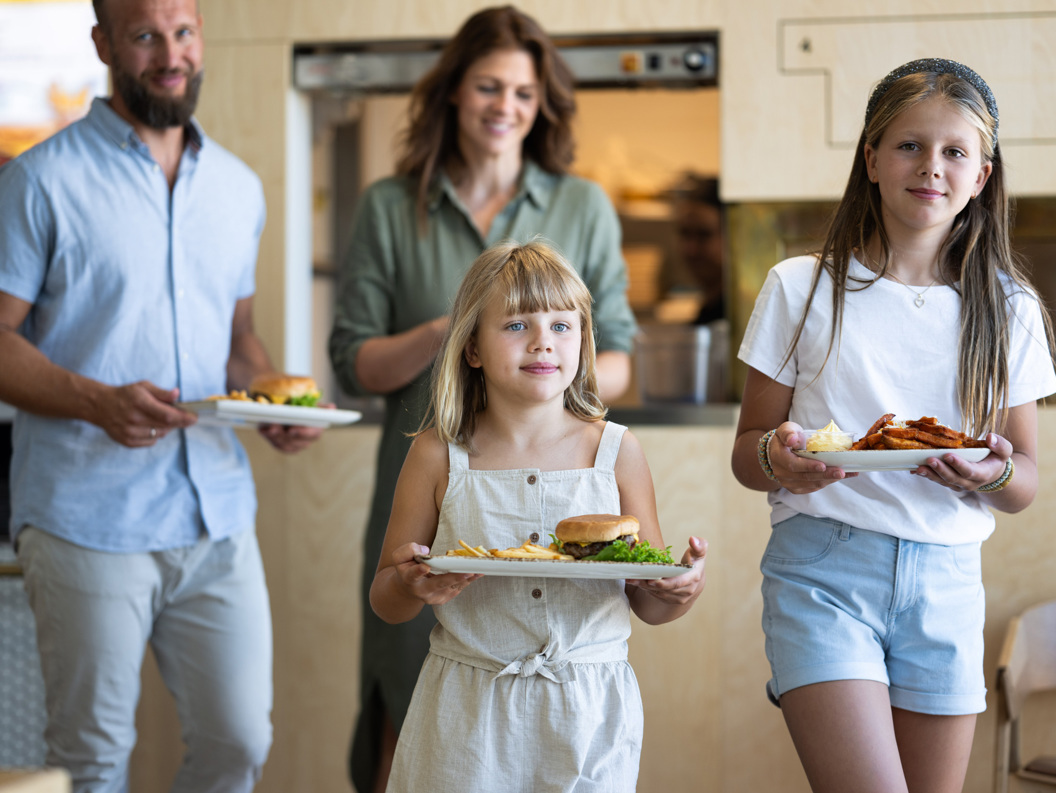 A family of four is served meals at a restaurant; father and mother in the background, two daughters in the foreground with burgers and fries on plates.