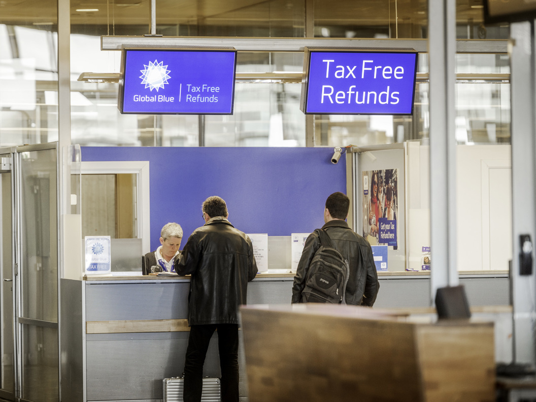 Personar ved ein Global Blue tax-free-refusjonsdisk på ein flyplass. To menn står ved skranken under eit blått skilt som viser "Tax Free Refunds".
