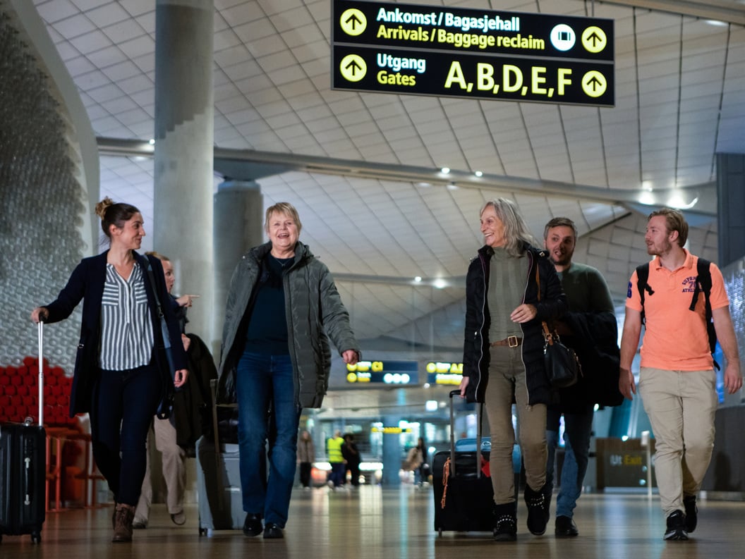Ei gruppe menneske går gjennom ein flyplass med bagasje, ankomst- og avgangsskilt synlege i bakgrunnen.