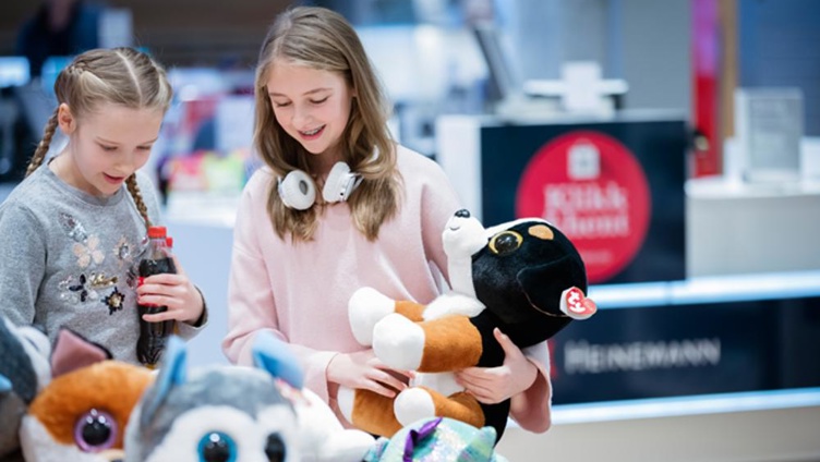 Two children are looking at stuffed animals in a store. One of them is holding a stuffed dog, while the other has a soda bottle.