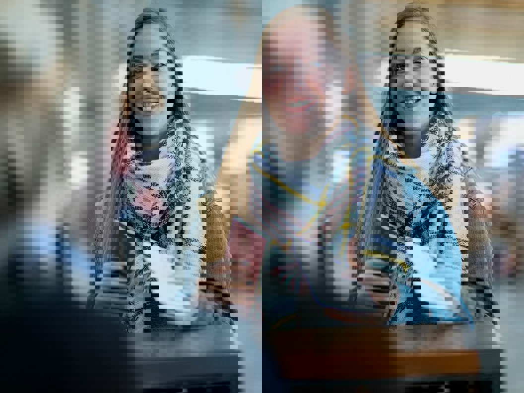 A lady with long, semi-dark hair is standing in front of a counter at the airport terminal with her passport in her hand.