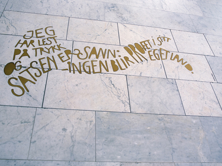 Tekst på gulv laget i gullbokstaver på marmorfliser, med teksten: "JEG HAR LEST PÅ TRYKK OG SAVN ER SATSEN: INGEN BLIR DRØFTET SÅ PROFT I SITT EGET LAND