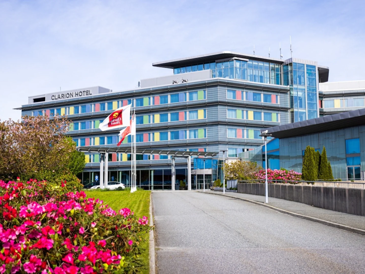 Moderne fasade av Clarion Hotel omgitt av frodige blomar og tre under ein klar himmel.