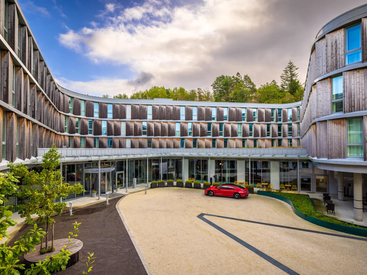 Moderne hotellfasade med bogeforma design, store vindauge, og ein raud bil parkert framfor hovudinngangen, omgitt av grøntområde og delvis overskya himmel.
