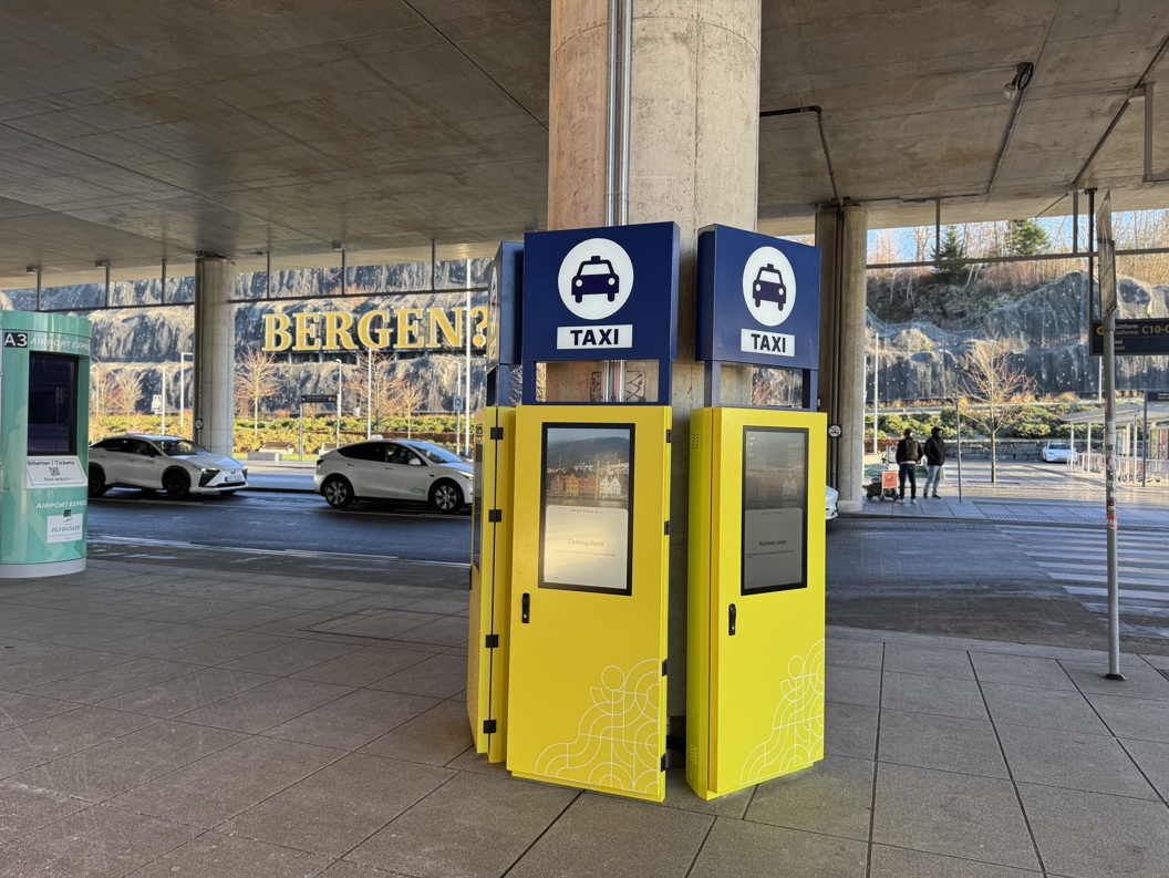 Taxiholdeplass på Bergen lufthavn med digitale kiosker og parkerte taxier, under en overbygd gangvei