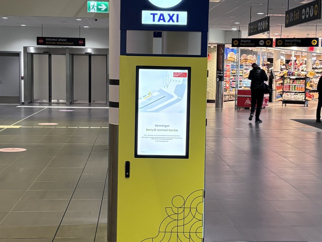 Taxisøyle på flyplass med skilt og digital skjerm for bestilling av taxi, plassert i nærhet av butikk og inngang