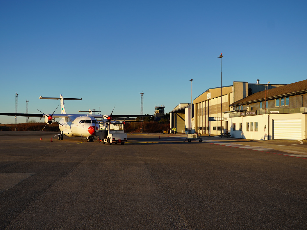 Widerøe-fly ved Florø lufthavn foran terminalbygningen, omgitt av skog og boliger i bakgrunnen.