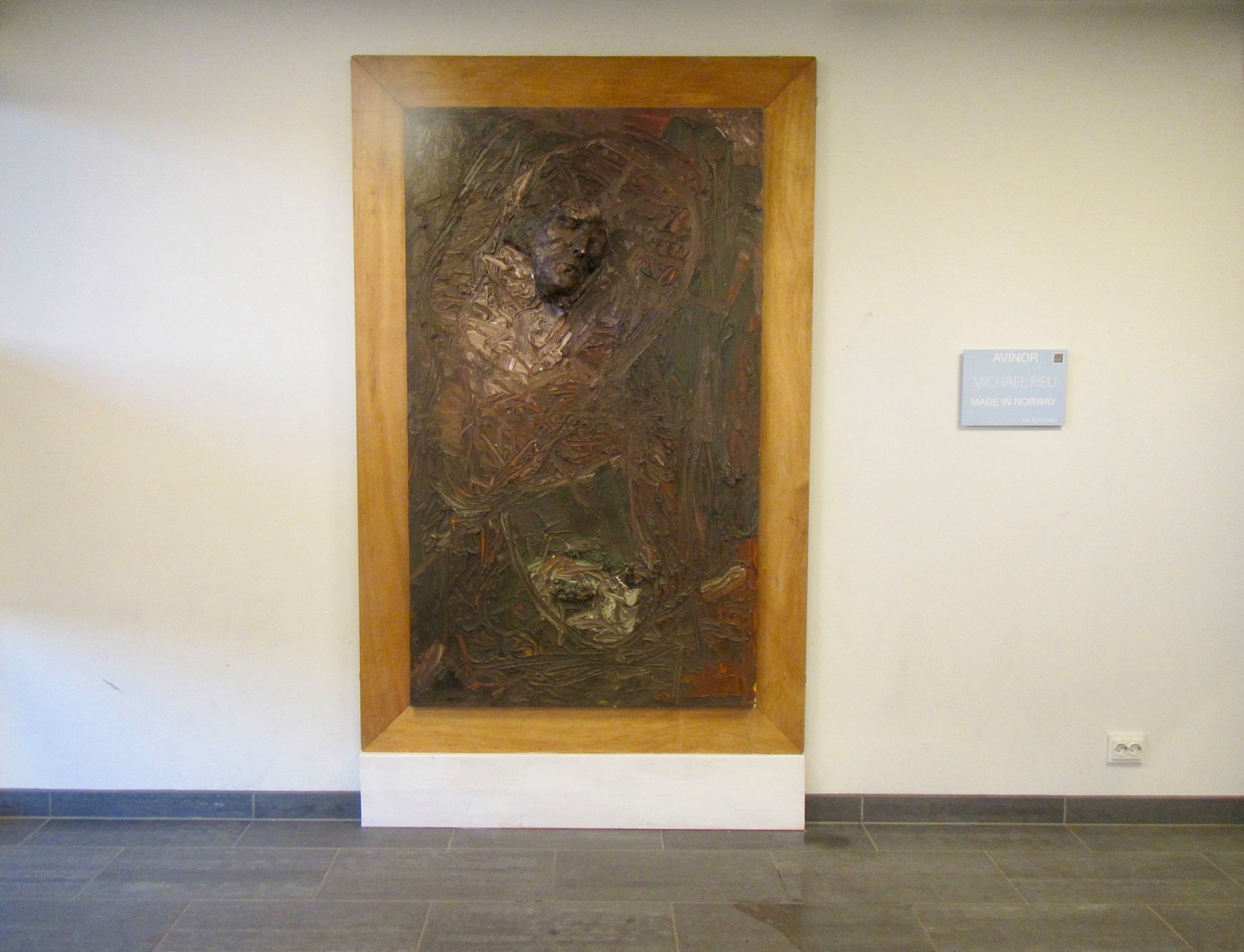 Vegghengt kunstverk i tre og metall, innrammet i en lystonet treramme, plassert i en lys gang