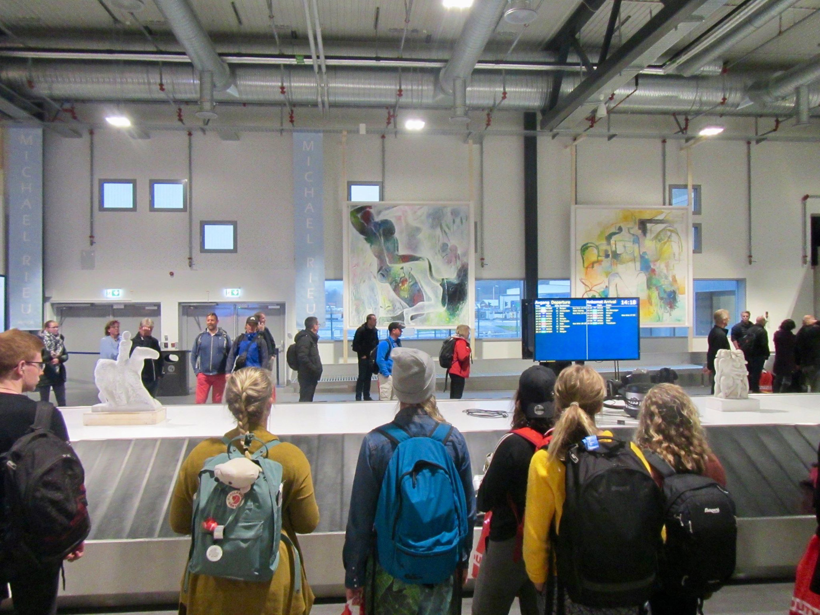 Passasjerer venter ved bagasjebåndet på en flyplass, med kunstverk på veggene i bakgrunnen