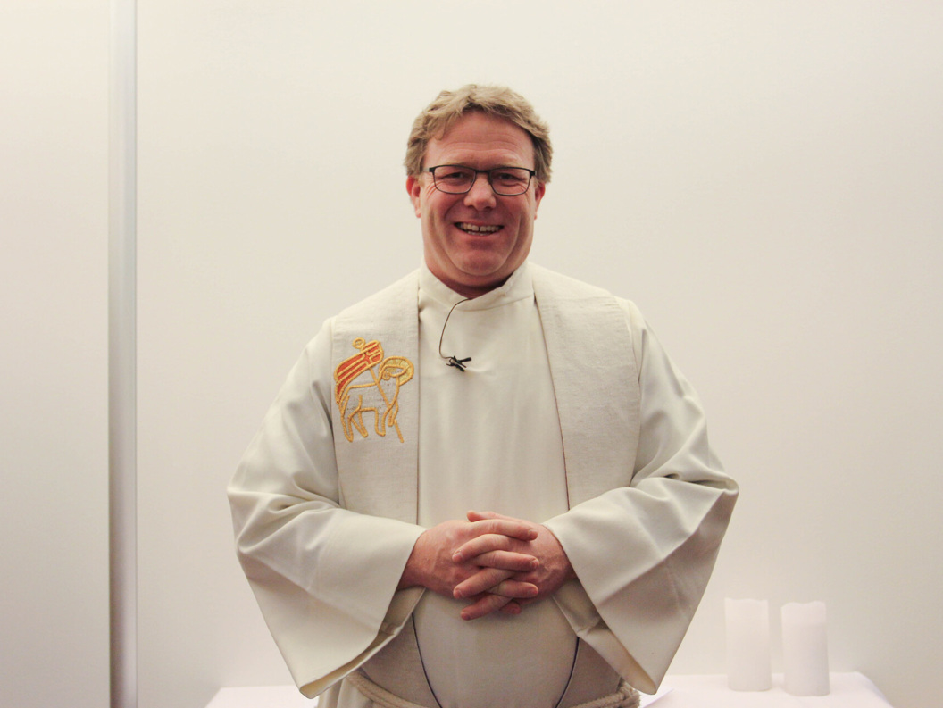 A smiling priest in a white robe stands in front of a light wall, with an embroidered symbol on the left side of the robe.