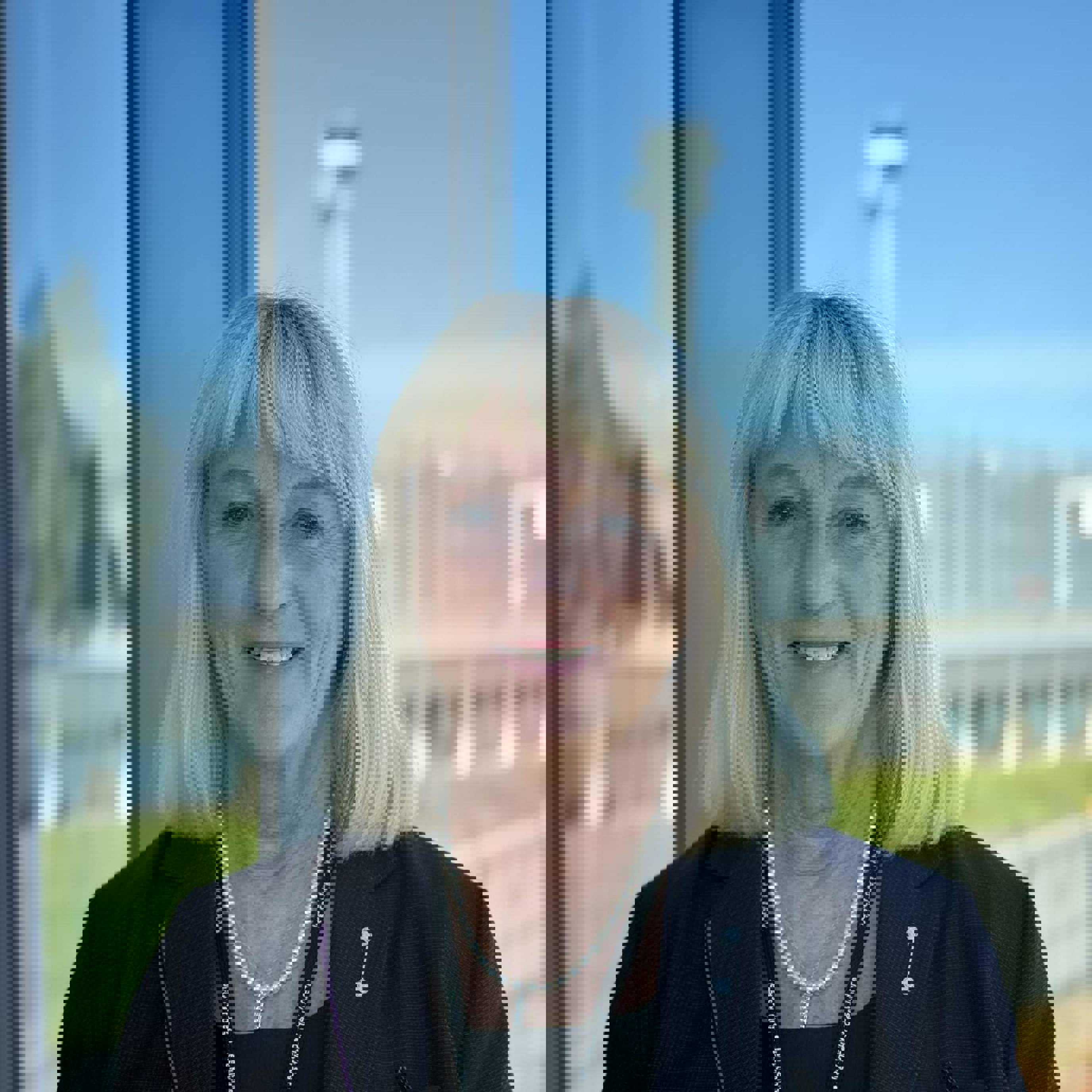 Woman with blonde hair in a dark blazer smiling in front of a window overlooking an open landscape and a blurred structure in the background.