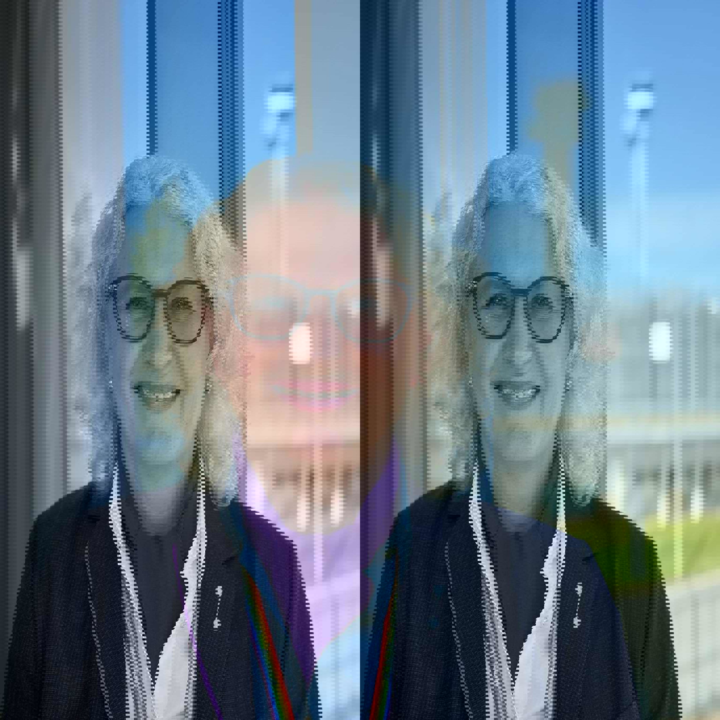 Woman with light curly hair and glasses stands by a window wearing a dark jacket and shirt, in front of an airport background with a clear blue sky.
