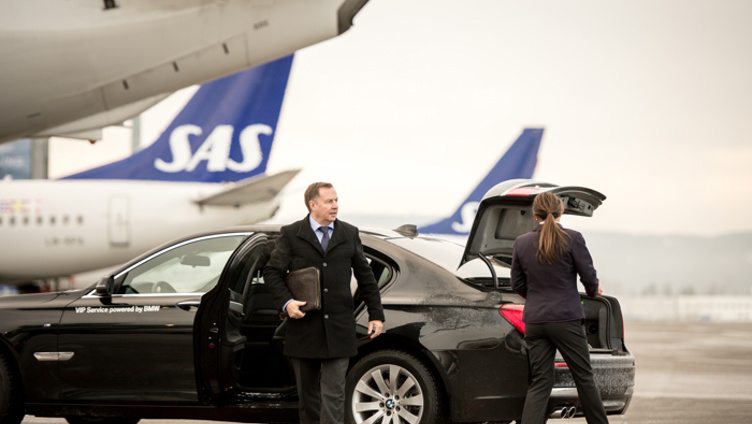 Businessman getting out of a luxury car at an airport, with SAS planes in the background. A woman is loading luggage into the car's trunk.