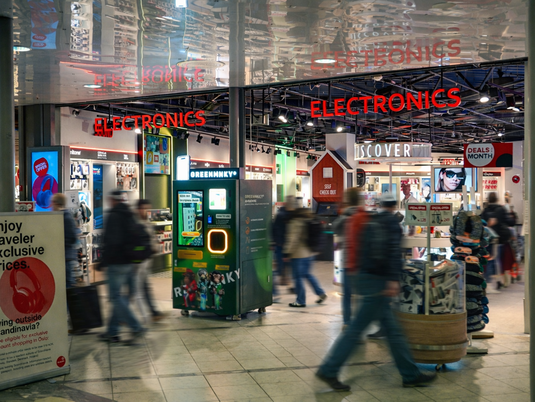 Selger som hjelper en kunde ved en CAPI-butikkdisk på en flyplass, omgitt av teknologiprodukter og fargerike hyller