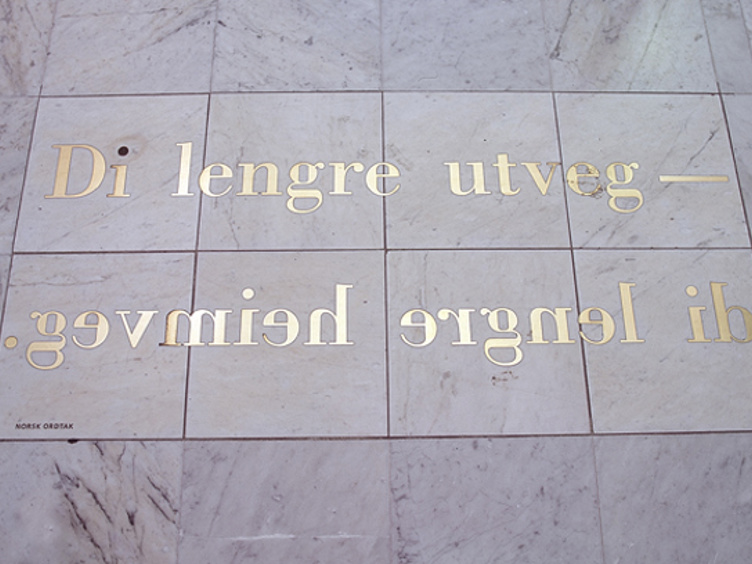 Gulletekst på et skilt montert opp-ned på en vegg med teksten "Di lengre utveg – di lengre heimveg" på norsk, designet av Kurt Johannessen. Skiltet er festet på en lys steinoverflate