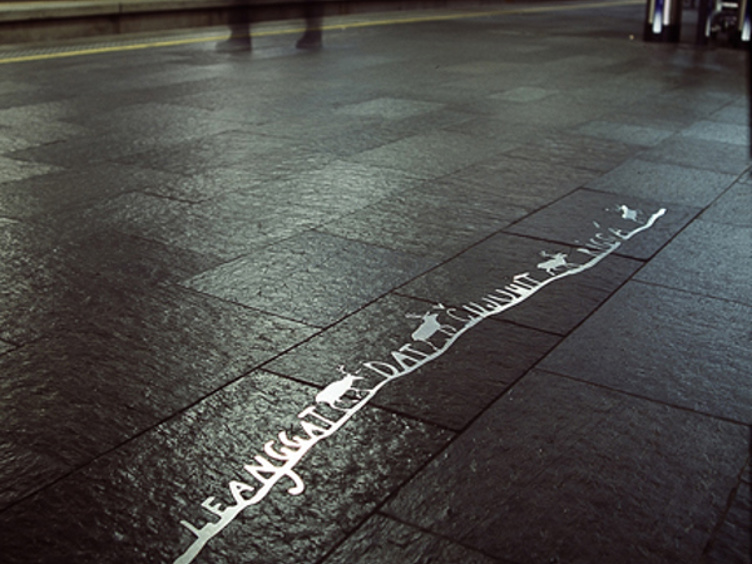 Utenfor et moderne bygg sees en gul strek med engelske instruksjoner på et fortau, omgitt av forbipasserende mennesker og automatporter i bakgrunnen