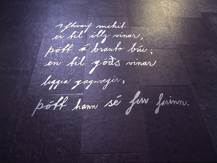 Gammelt sitat skrevet på mørkt gulv i gotisk skrifttype, opplyst av svakt lys. Teksten handler om vennskap og vises mot en steinlignende bakgrunn