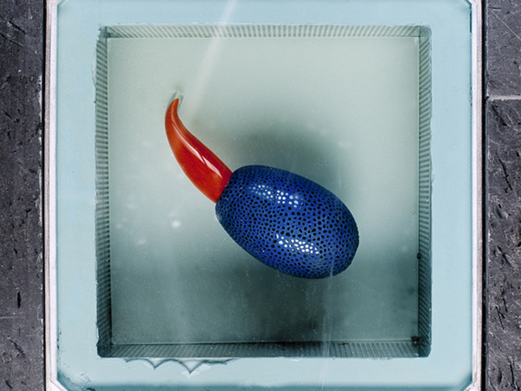 Blå skulptur med rødt utspring sett ovenfra, plassert i en glassinnrammet åpning