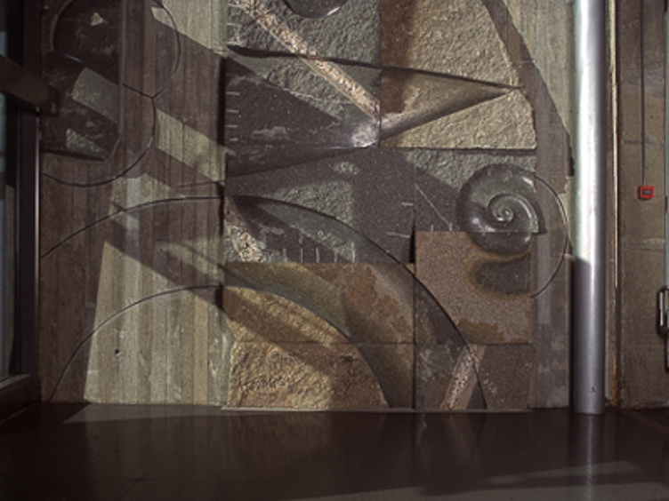 Veggfreske i stein med abstrakte former og geometriske mønstre, opplyst av naturlig lys fra en glassvegg på en moderne bygning