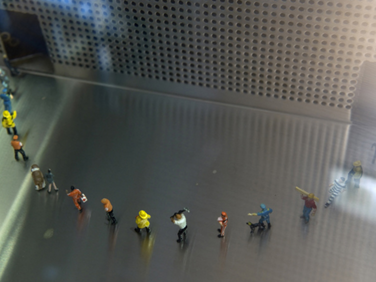 Miniatyrfigurer i kø på metalloverflate ved perforert bakgrunn, med variert bekledning og farger