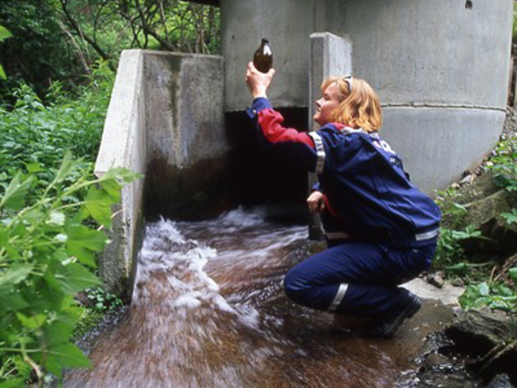 Avinor employee taking a water sample in a stream, wearing a blue rain jacket, surrounded by green vegetation.