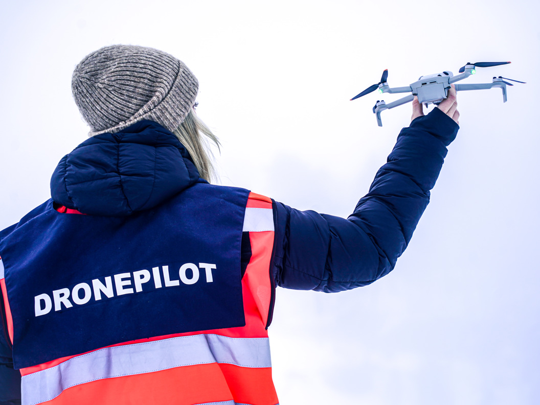 Kvinne i oransje jakke med dronepilot-vest som holder en drone i hånden opp mot himmelen