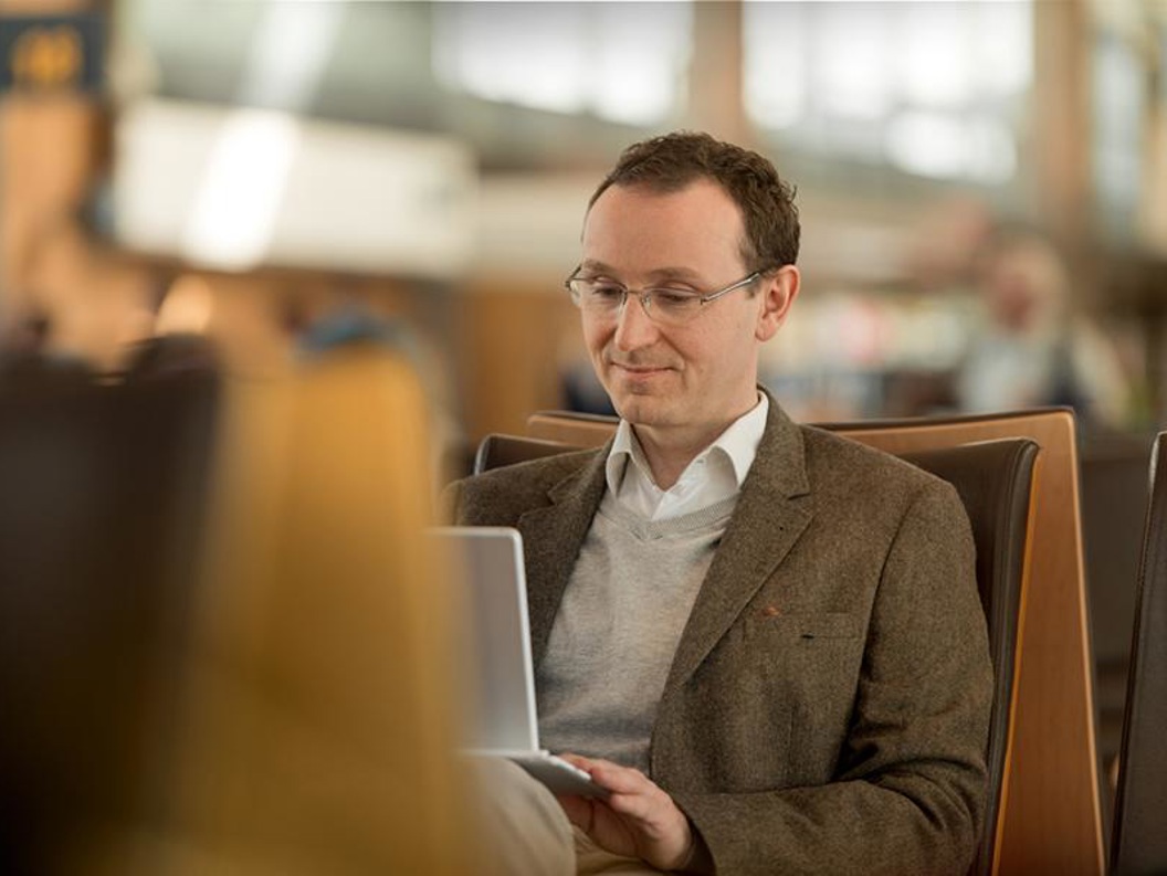 Ein mann med kort brunt hår og briller som sit på ein av plassane ved ein gate og ser på den bærbare pc-en sin som han har på fanget.